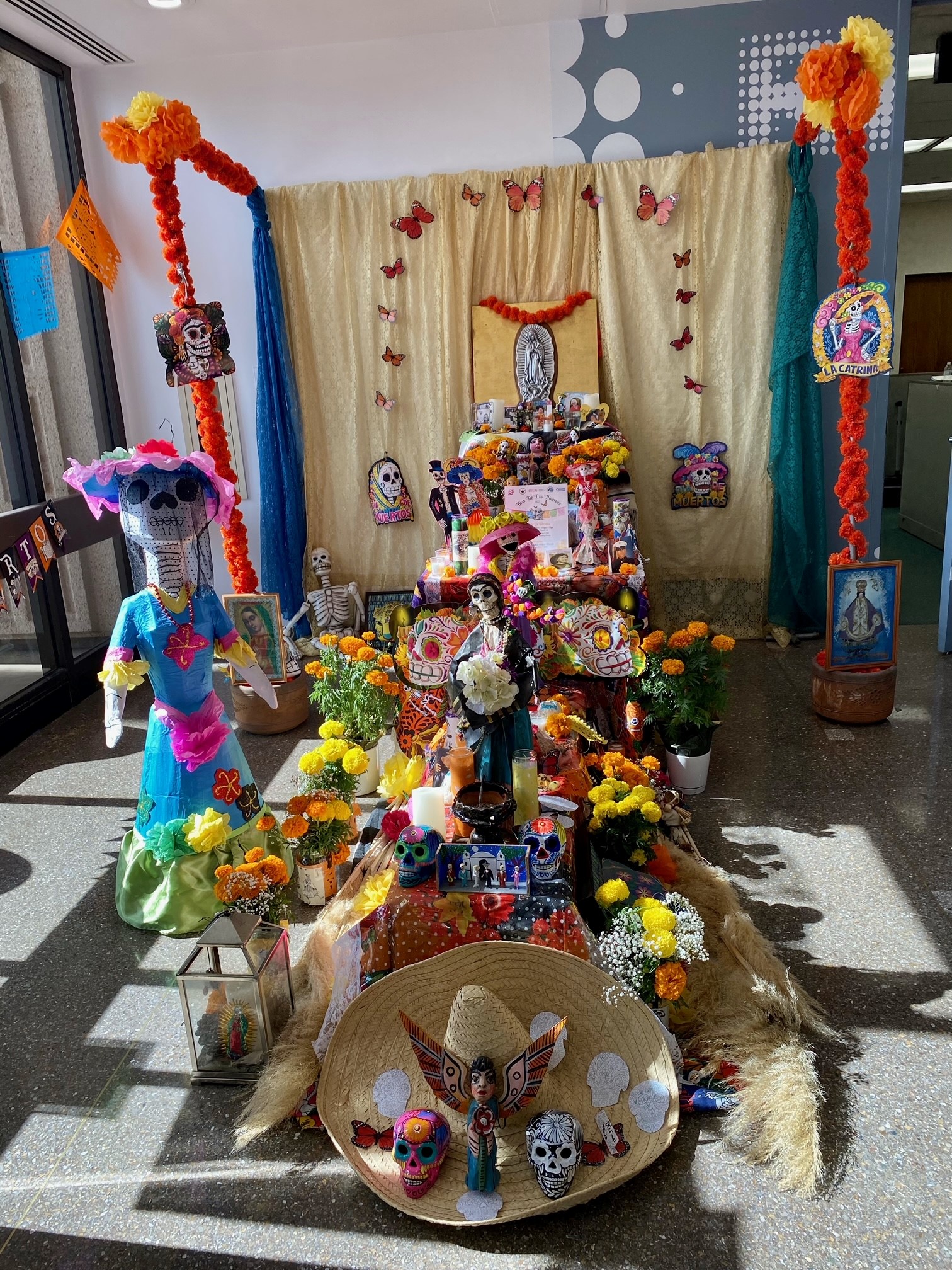A colorful dia de los muertos altar, framed by orange marigolds, a paper mache Catrina figure, and monarch butterflies