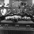 Men form a chain of clasped hands behind the casket of Dr. Martin Luther King Jr. during his eulogy at Morehouse College. Left to right on the podium: Dr. Ralph David Abernathy (background, behind lectern, top of head visible only), Fred Shuttlesworth (background, right of lectern), Benjamin Hooks (right of Shuttlesworth). Front row left to right: unidentified African American man, Leon Hall, Al Sampson, Cirillo McSween, Y.T. Rogers, J.T. Johnson, James Bevel, and two unidentified African American men. 1968