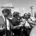 Civil rights advocate James Meredith appears to be doing a media interview. Occasion was the James Meredith march for civil rights and voter registration. In background men and women display protest signs with civil rights/voter registration messages. 1966, Harry Adams. 93.01.HA.N120.B9.12.84.08 