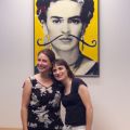Two women in front of a picture of Frida Kahlo with a mustache