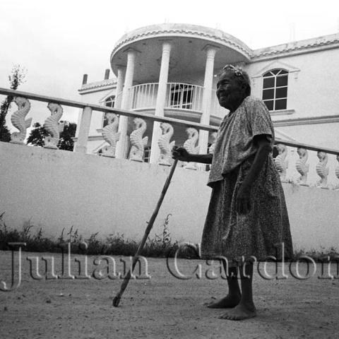 An elderly woman with a walking stick walking in front of a mansion