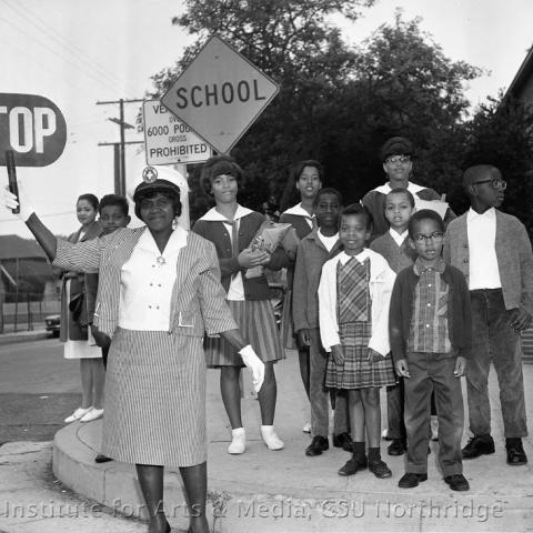 Woman holding a stop sign and kids waiting on the sidewalk.