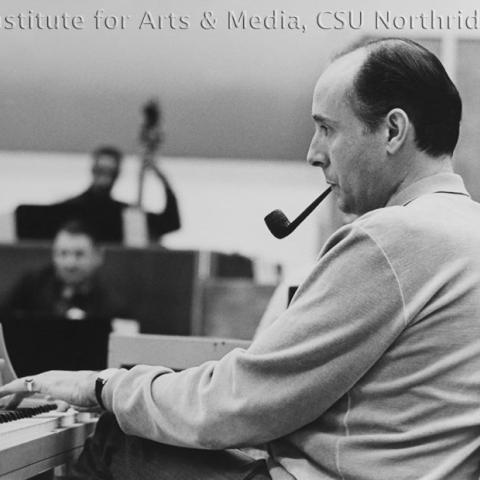 Man sits in front of a piano with a tobacco pipe in his mouth.