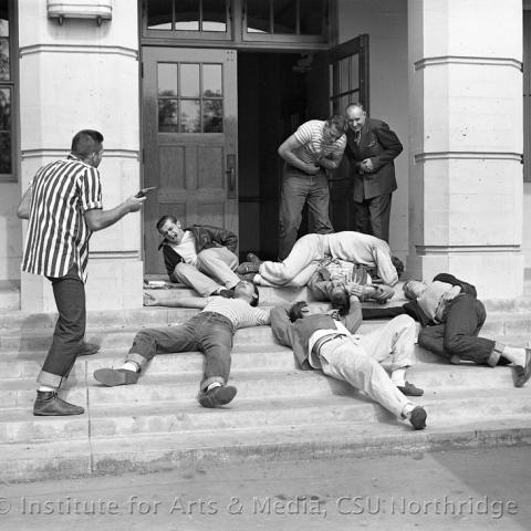 People act dead at the entrance of a building while another man holds a gun.