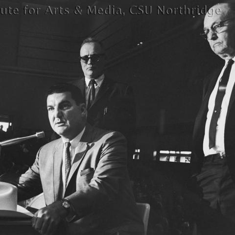 A man speaking over the microphone while two men stand behind him.