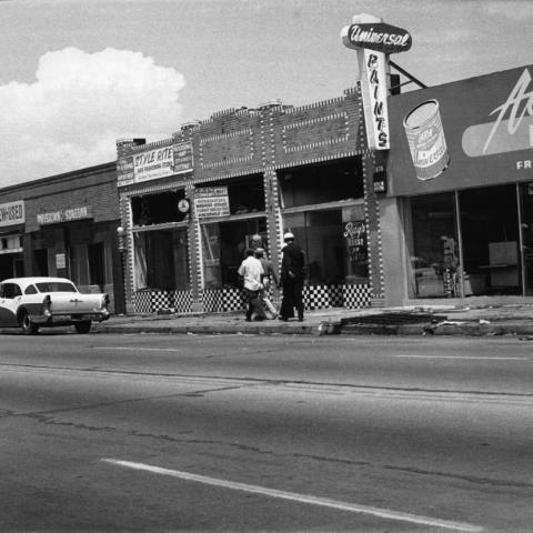Police officers and two African American men walk along a sidewalk in front of damaged storefronts in the aftermath of the Watts Riots, 1965, Guy Crowder Collection, Tom & Ethel Bradley Center Photographs. 