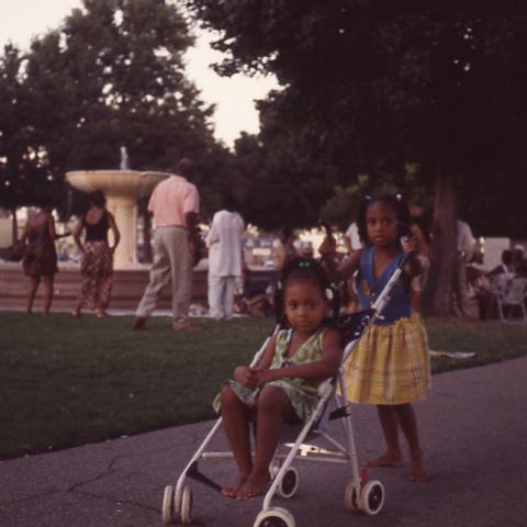 A young girl pushes a younger girl in a stroller near the Leimert Park fountain, 1998. Roland Charles Collection, Box 75.
