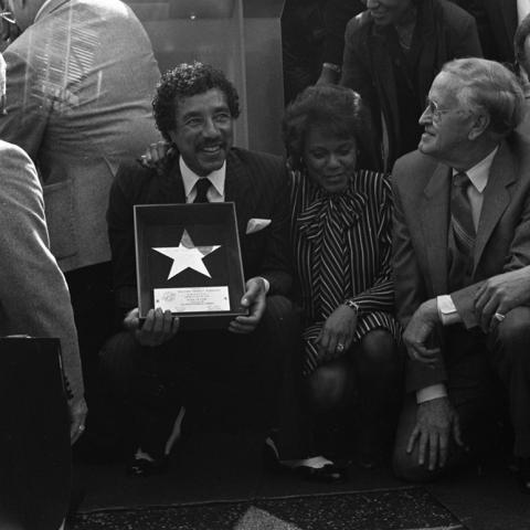 Smokey Robinson kneels, holding a framed commemoration, near his star on the Hollywood Walk of Fame as Claudette Robinson (2nd from right) and an unidentified man kneel next to him while a crowd is looking on. Guy Crowder Collection
