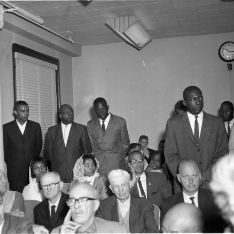 Several men in suits stand around a seated Malcolm X, Harry Adams Photograph Collection