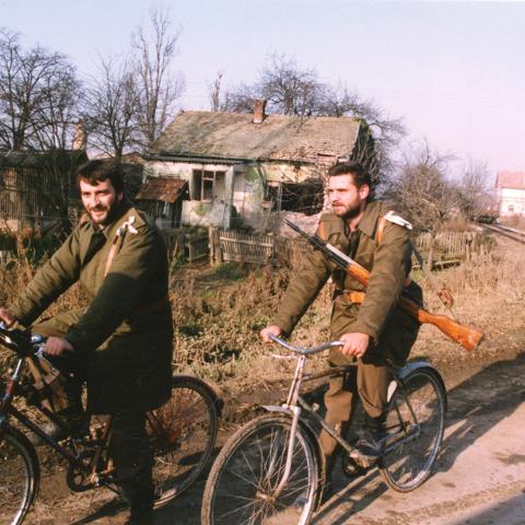 Two Croatian troops on bicycles riding past a dilapidated house in the midst of war. ca. 1991-1994. Box 1, Folder 12. 