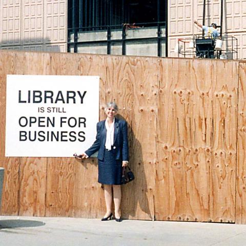 Library Dean, Susan Curzon, poses next to a sign that reads "Library is still Open for Business"