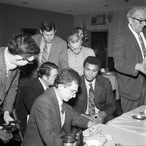 Heavyweight Champion Muhammad Ali surrounded by journalists and media personal for a press conference to discuss his future computer fight with Heavyweight champion Rocky Marciano, 1970. Harry Adams Collection. ID: 93.01.HA.N120.B8.38.89.02