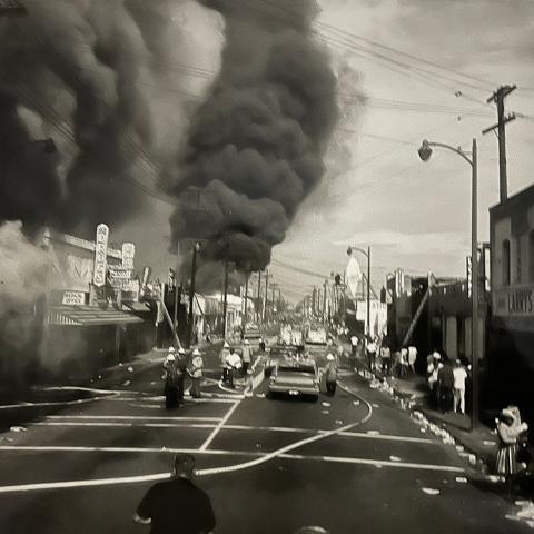 A view down a city street in Watts with dark smoke billowing up in the distance. Fire personnel, a car and fire engines are in the street and residents are standing on the sidewalks, 1965, League of Women Voters of Los Angeles Collection. 