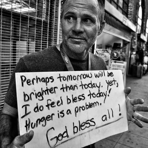 Portrait of Patrick holding a sign that reads, “Perhaps tomorrow will be brighter than today. Yet, I do feel Bless [sic] today! Hunger is a problem. God bless all!”, ca. 1990.