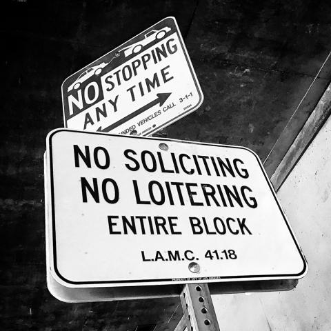 Street signs intended to keep the area clear of people. The signs read, “NO STOPPING ANY TIME” and “NO SOLICITING NO LOITERING ENTIRE BLOCK L.A.M.C. 41.18”, ca. 1990.
