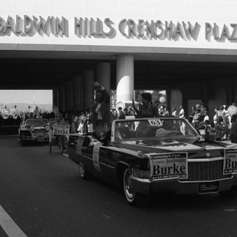 Yvonne Brathwaite Burke waving from a limousine convertible decorated with posters from her campaign for Los Angeles County Supervisor in the Kingdom Day parade. A woman on a tricycle follows the limousine as the parade route passes Baldwin Hills Crenshaw Plaza. 1992. Digital ID: 11.06.GC.N35.B5.47.158.10A