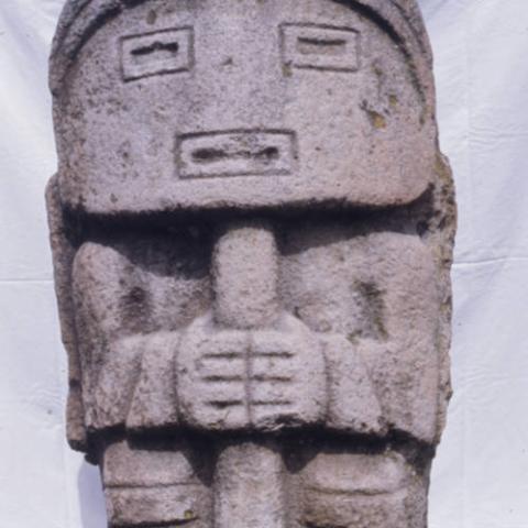 Stone statue of a figure with a mask-like face and holding a staff stands at the Ullumbe site in San Agustín Archaeological Park. Photo taken in 1975. Digital ID: 99.01.RCr.sl.B17.04.28.05