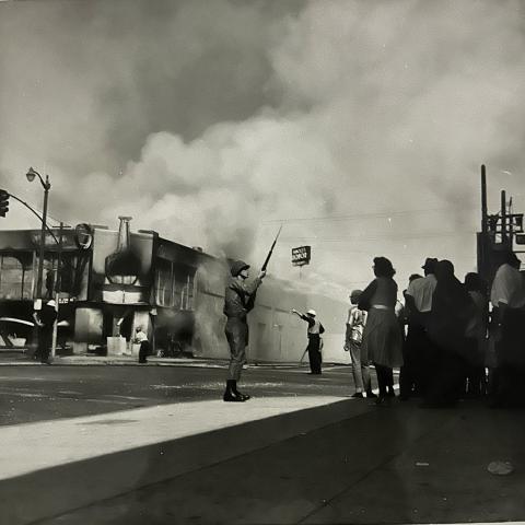 A National Guard member with a rifle and bayonet stands in the street in front of a crowd in Watts. A burned-out building is behind him and smoke fills the sky, 1965, League of Women Voters of Los Angeles Collection. 