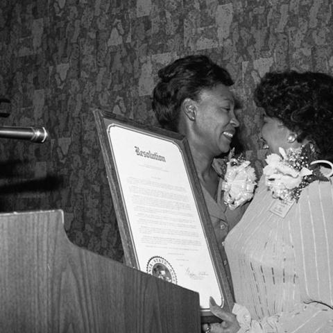State Assembly member Maxine Waters stands at a lectern with an unidentified woman as she presents a framed copy of a resolution to a representative of the National Association of Media Women during their 18th annual convention hosted this year by the Beverly Hills/Hollywood Chapter. Ms. Waters was guest speaker during the President's luncheon. 1983. Digital ID: 11.06.GC.N35.B19.58.96.15