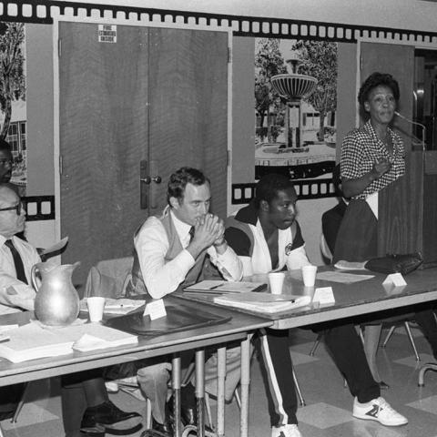 Maxine Waters speaks at a lectern next to a panel of people seated at a table during a community meeting on jobs. 1984. Digital ID: 11.06.GC.N35.B11.10.13.35A
