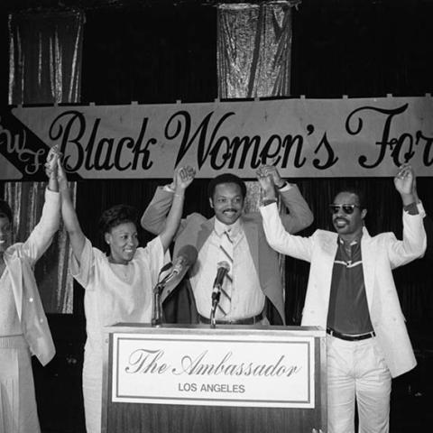 Maxine Waters (2nd from left), Jesse Jackson (center) and Stevie Wonder (2nd from right) stand at a lectern with raised clasped hands between an unidentified man and woman during a Black Women's Forum event at the Ambassador Hotel. Digital ID: 11.06.GC.N35.B6.21.59.10A