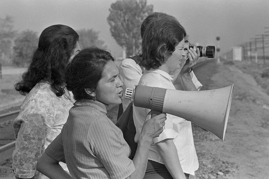 Dolores Huerta speaking through a megaphone, standing with other women in a field.