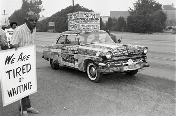 Man holds picketing sign that says 'WE ARE TIRED OF WAITING' next to car covered with protest stickers