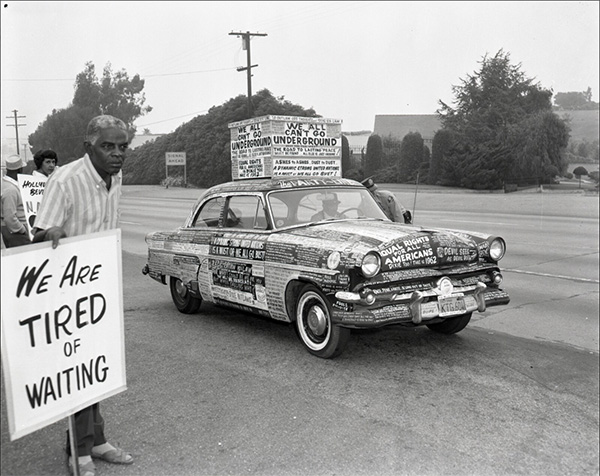 Man holds picketing sign that says 'WE ARE TIRED OF WAITING' next to car covered with protest stickers