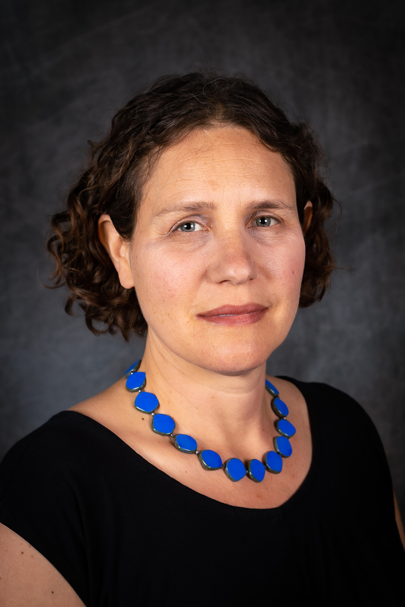 Profile of Daniela Gerson wearing a black dress and blue beaded necklace amid a dark grey background