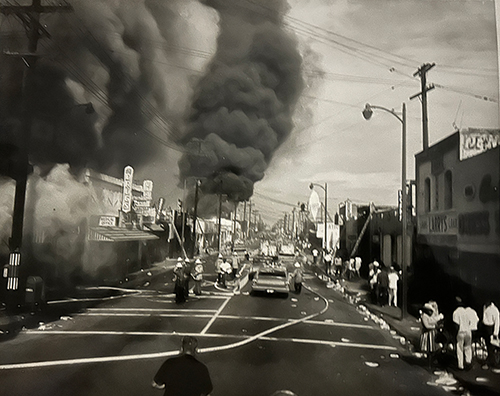 A view down a city street in Watts with dark smoke billowing up in the distance, 1965.