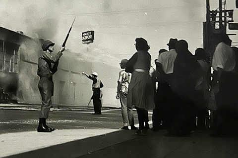 A National Guard member with a rifle and bayonet stands in the street in front of a crowd in Watts. A burned-out building is behind him and smoke fills the sky, 1965, League of Women Voters of Los Angeles Collection. 