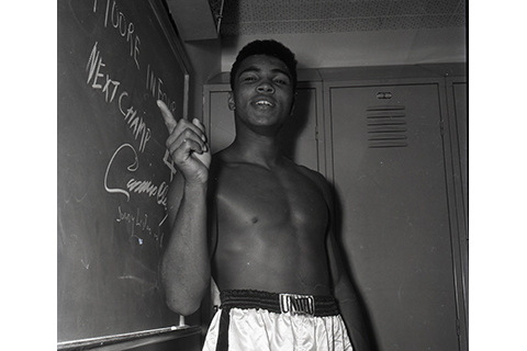 Cassius Clay, (soon to be Muhammad Ali) points to a chalk board that reads "Moore in 4, Next Champ Cassius Clay" at an event leading up to his boxing match against Archie Moore at the Sports Arena, November 15, 1962,  Harry Adams Collection. ID: 93.01.HA.B4.N120.605.1