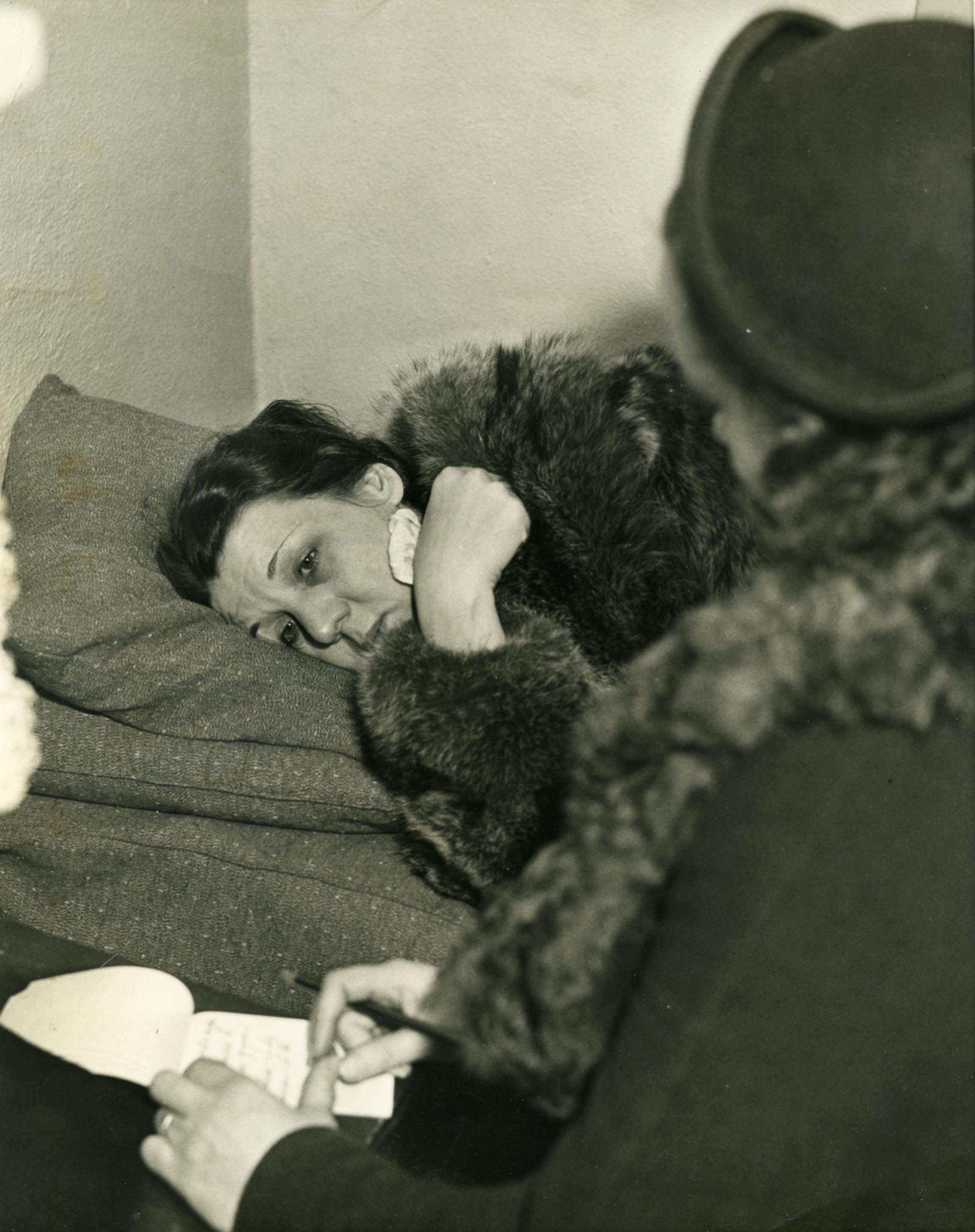 Helen Wills lies on a couch in a fur coat, looking dazed. A reporter sits in the foreground with a notepad.