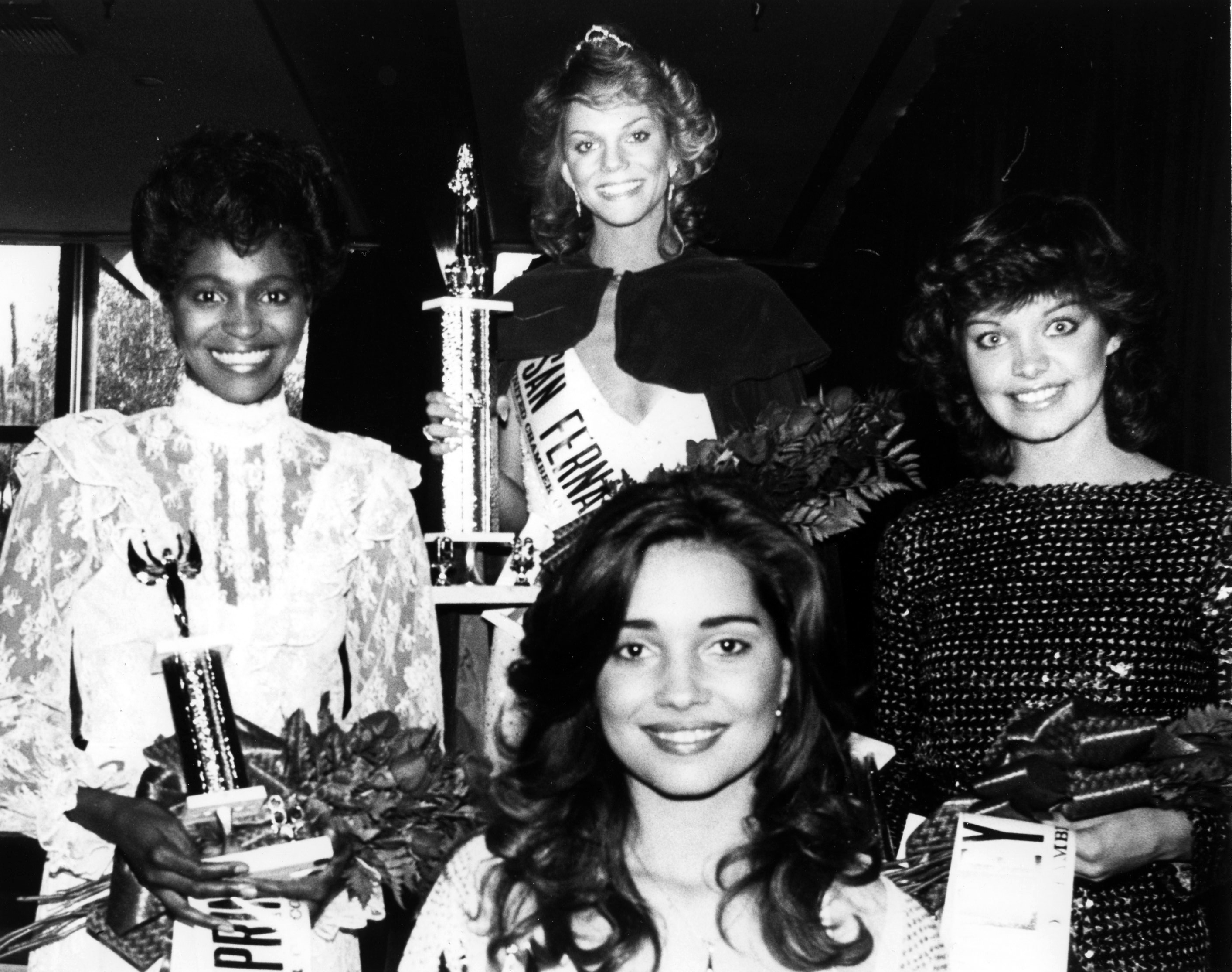 A diverse group of four women; the pageant queen wears a crown, with the three runners up, holding trophies and bouquets.