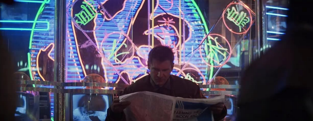 Man sitting down reading newspaper. In background, various colorful neon designs and lights adorn the window glass behind him.