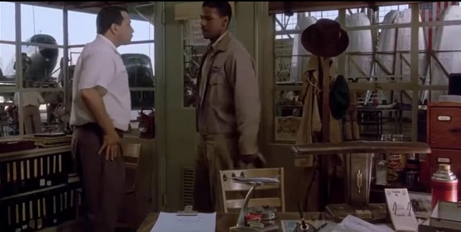 Two men, one black and the other white, talking inside the office of an airplane hangar.